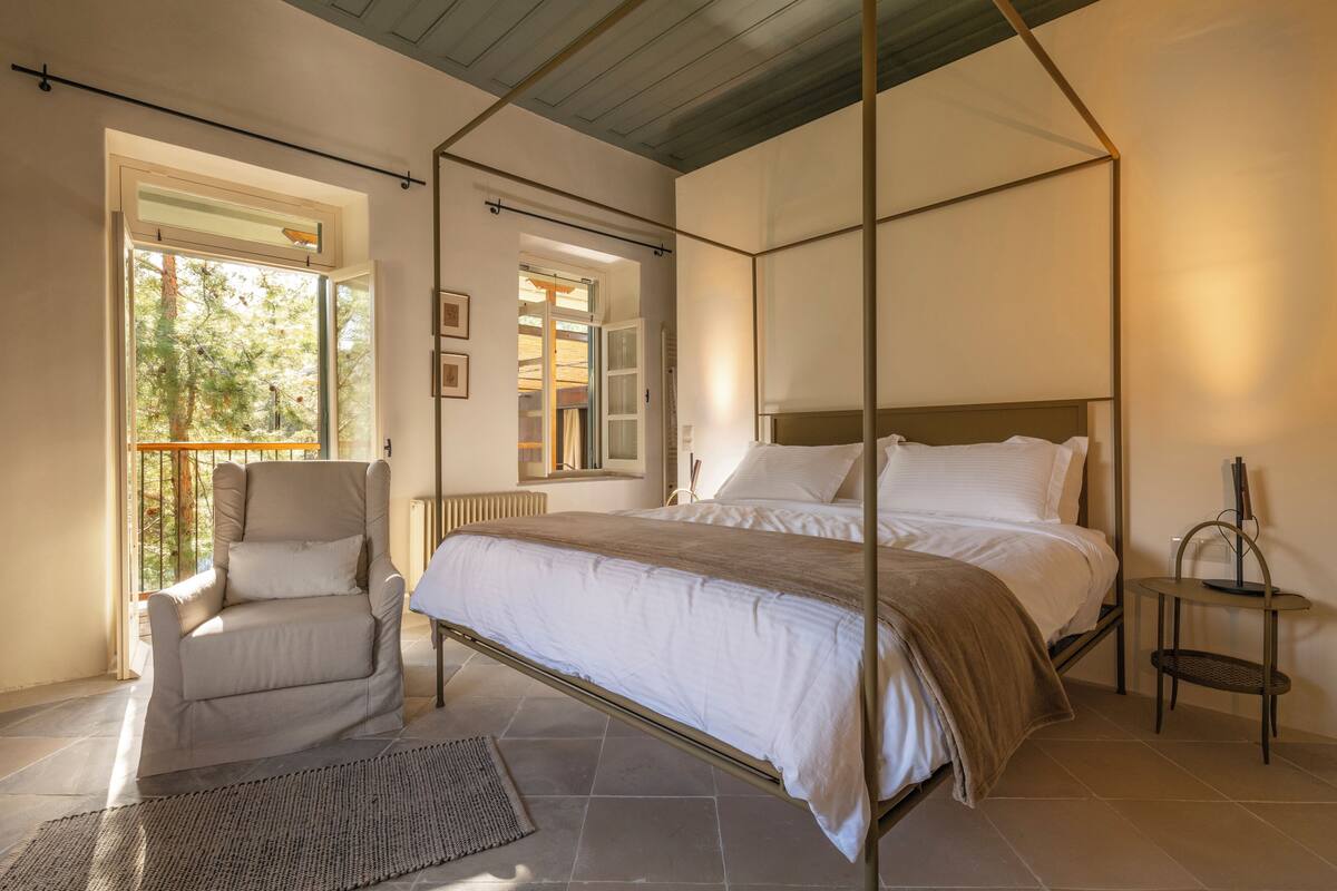 A bedroom with a four-poster bed, a chair, a small table, and a view of the outdoors.