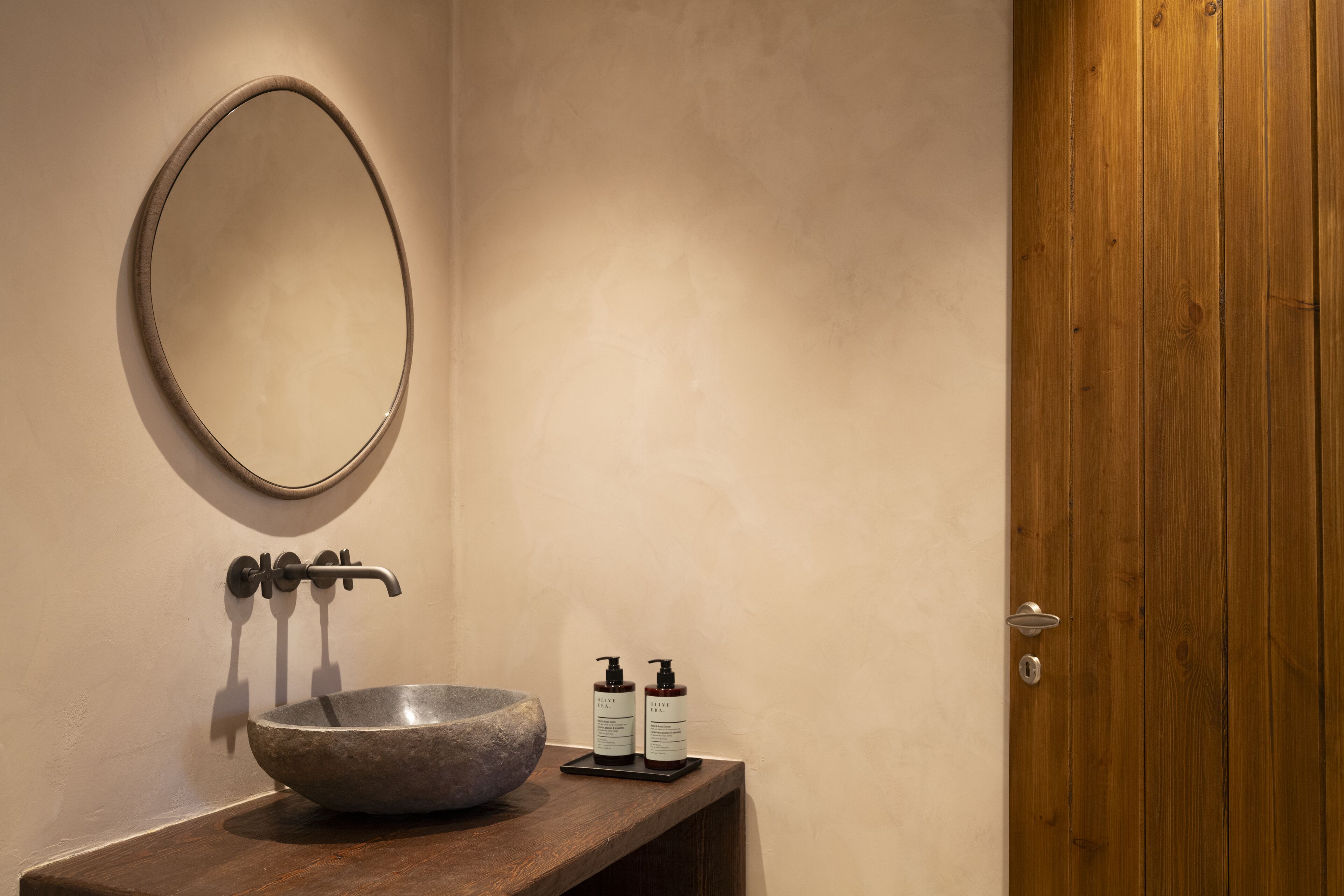 A bathroom with a stone sink, a round mirror, a wooden door, and two bottles of shampoo.