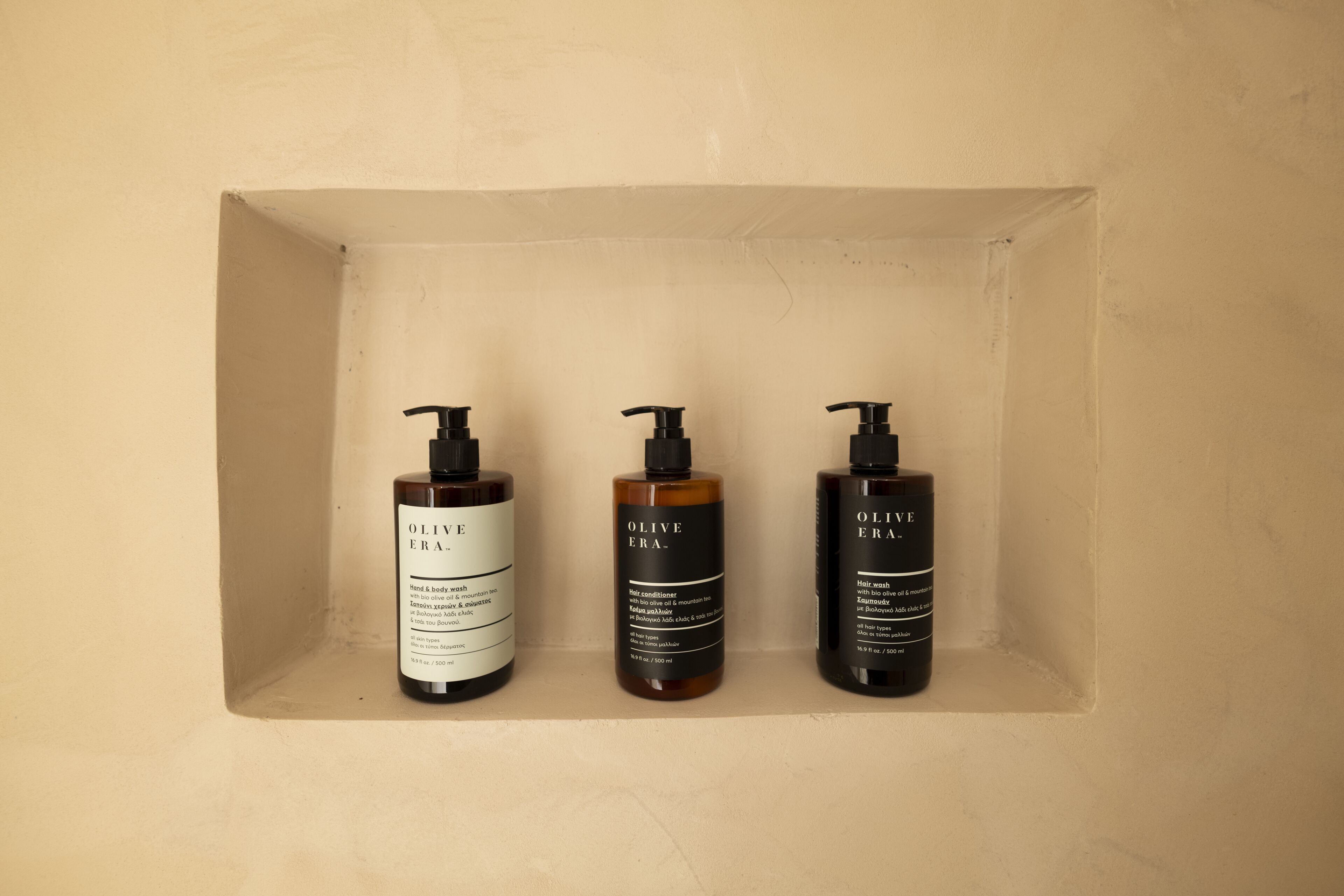 Trois bouteilles de gel douche et shampooing Olive Era sont posées sur une étagère.