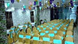 A banquet hall with rows of chairs covered in gold fabric, a green carpet, and colorful balloons. - Way Inn Familiar (Hyderabad)