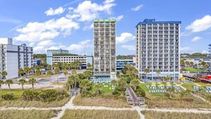 A high-rise hotel with multiple floors, a parking area, and a landscaped courtyard with palm trees. - King Suite, Kitchen, New HVAC, Ocean Views, Walk to Attractions & Restaurants (Myrtle Beach)