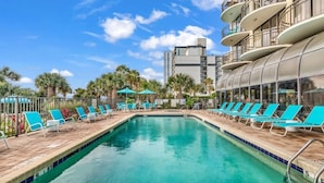 A swimming pool with lounge chairs and umbrellas, surrounded by palm trees and a building with balconies. - King Suite, Kitchen, New HVAC, Ocean Views, Walk to Attractions & Restaurants (Myrtle Beach)
