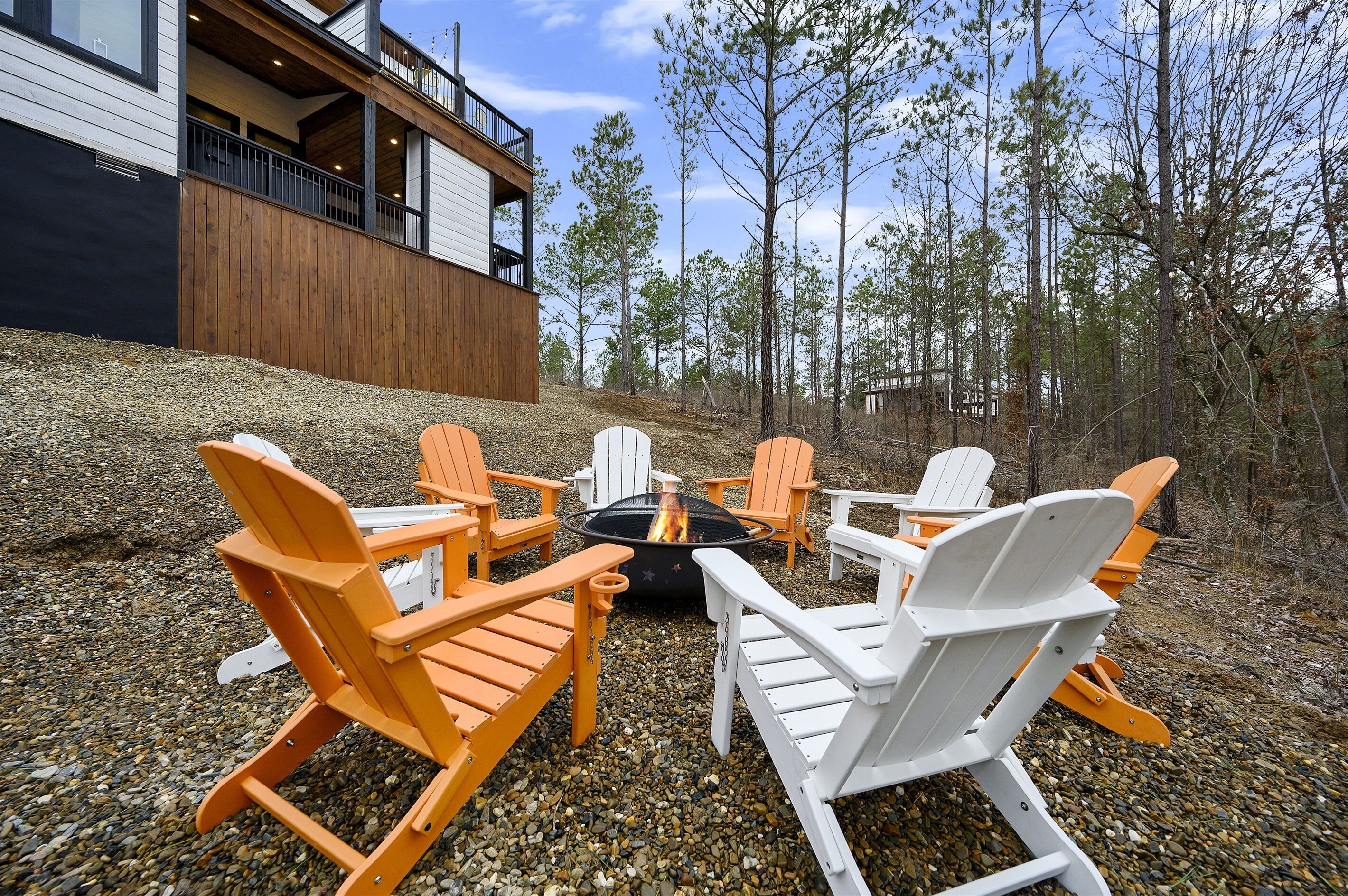 A deck with Adirondack chairs and a fire pit surrounded by a gravel area, with a building in the background.