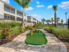 A golf putting green with a hole marked, surrounded by a landscaped area with palm trees and a building in the background.