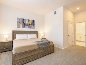 A hotel room with a wooden bed, two bedside tables, a painting on the wall, and a bathroom visible through an open door.