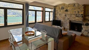 A living room with a stone fireplace, a glass dining table, and large windows overlooking a scenic view. - Riflessi sul Lago Apt Laglio - Tramonto (Laglio)