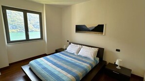 A bedroom with a bed, a window with a view, a nightstand, and a wall-mounted picture. - Riflessi sul Lago Apt Laglio - Tramonto (Laglio)