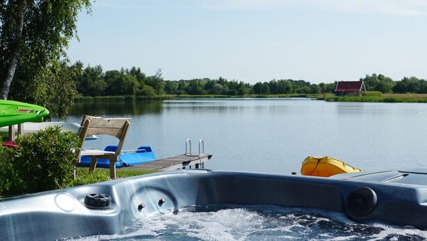 A hot tub with a wooden deck, surrounded by a calm lake, a red-roofed building in the distance, and lush greenery. - Lieskehuis Wellness de Luxe 10+2 by Interhome (Vlagtwedde)
