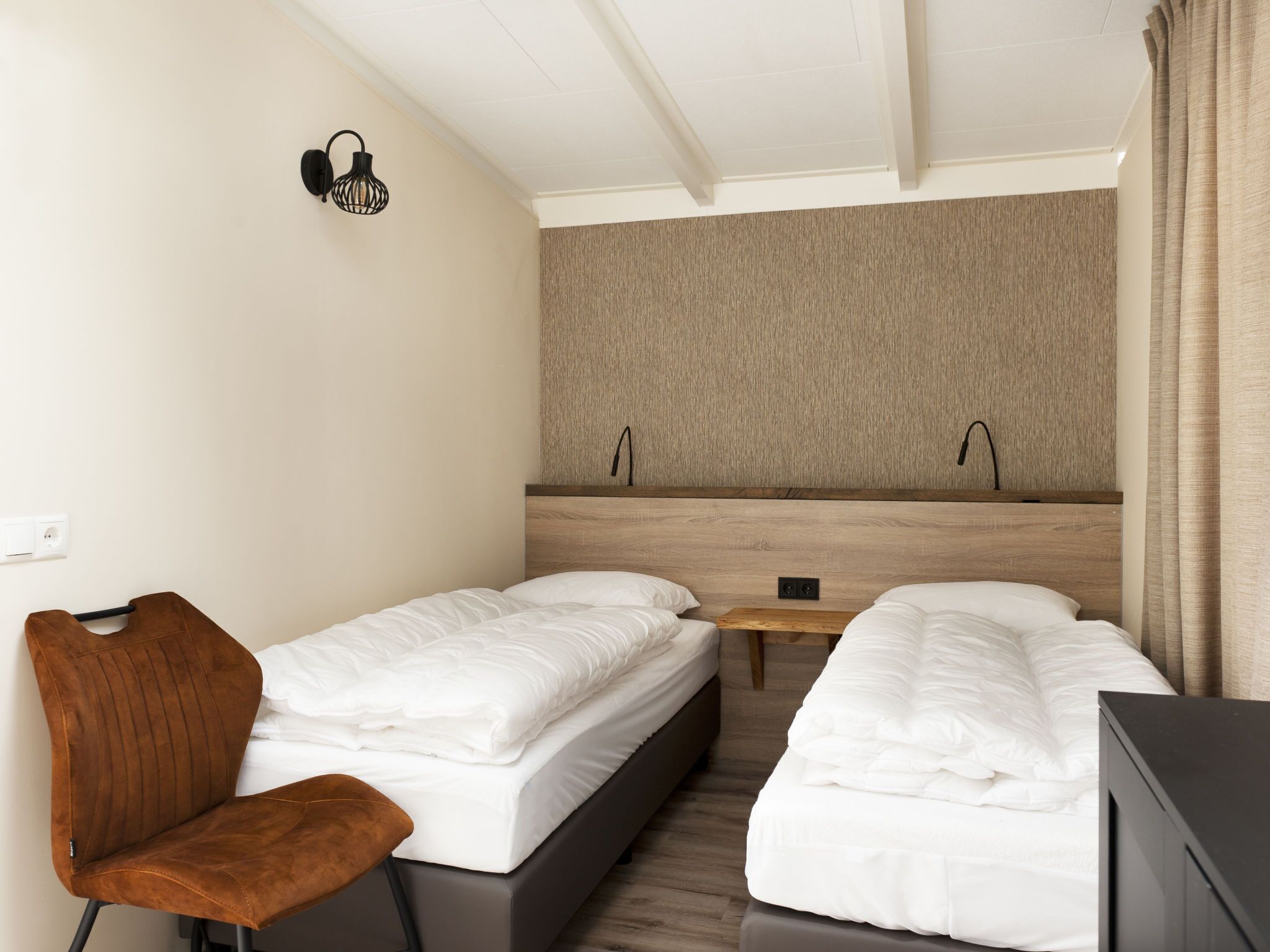 Hotelkamer met twee enkele bedden, een klein houten tafeltje, een bruine stoel, een wandlamp en een effen wand met een structuurpaneel.