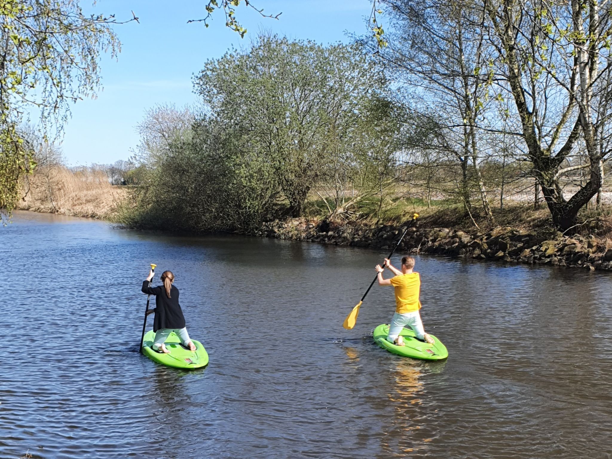 Deux personnes sur des planches de paddle vertes, sur une rivière paisible bordée d’arbres.