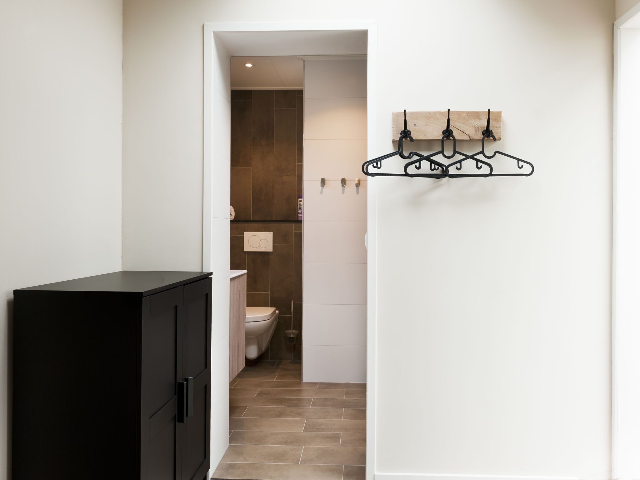 A narrow hallway with a black cabinet on the left and a bathroom with a toilet and shower visible through an open door.