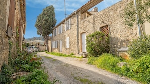 Une ruelle étroite pavée, bordée de bâtiments en pierre dotés de portes et de fenêtres en arc de cercle, entourée de verdure et baignée d’un ciel dégagé.