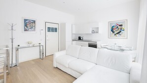 A modern, white-themed living room with a white sofa, a glass coffee table, and a kitchen area with white cabinets and a black countertop.