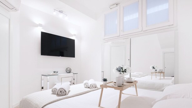 A modern hotel room with a large bed, a bedside table with a tray, a flat-screen TV mounted on the wall, and a large mirror reflecting the room.