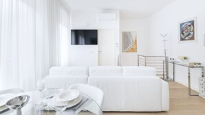 A modern, white-themed living room with a sofa, a glass table with wine glasses and plates, and a staircase leading to an upper level.