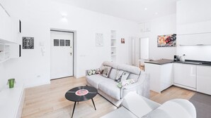 A modern, white-themed living room with a sofa, a coffee table, and a kitchen area with white cabinets and a countertop.