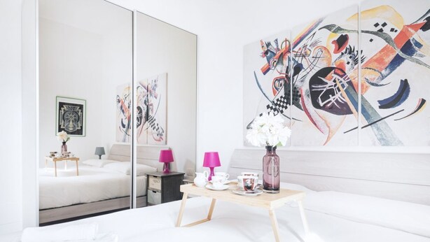 A modern hotel room with a large mirror, a bed, a small table with tea set, and abstract art on the wall.