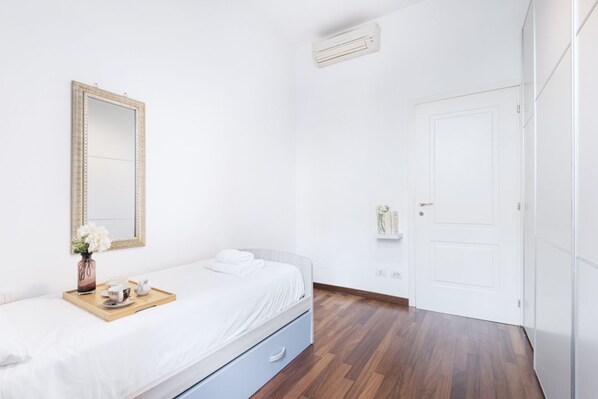 A bedroom with a bed, a bedside table with a tray, a mirror, a door, and an air conditioning unit.