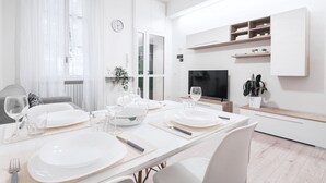 Un espace repas moderne avec une longue table dressée pour quatre personnes, des chaises blanches et un téléviseur fixé au mur.