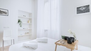 Une chambre d’hôtel moderne avec un grand lit, une table de chevet avec un plateau, une chaise, une armoire blanche et une fenêtre avec des rideaux légers.