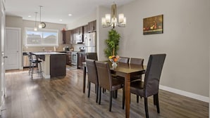 Une cuisine moderne avec des meubles en bois foncé, des appareils électroménagers en acier inoxydable et un îlot central. On y trouve un coin repas avec une table et des chaises en bois, ainsi qu’une grande fenêtre offrant une vue sur l’extérieur.