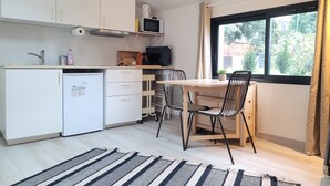 Une cuisine compacte avec des meubles blancs, un four à micro-ondes et un petit réfrigérateur. On y trouve une table à manger avec deux chaises et une fenêtre donnant sur un espace verdoyant.