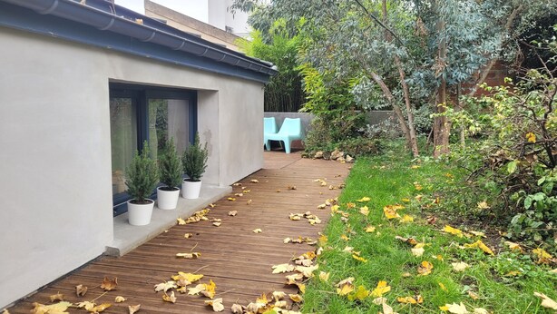 Un bâtiment moderne doté d’une terrasse en bois, de plantes en pot et d’un espace jardin avec de la pelouse et des arbres.