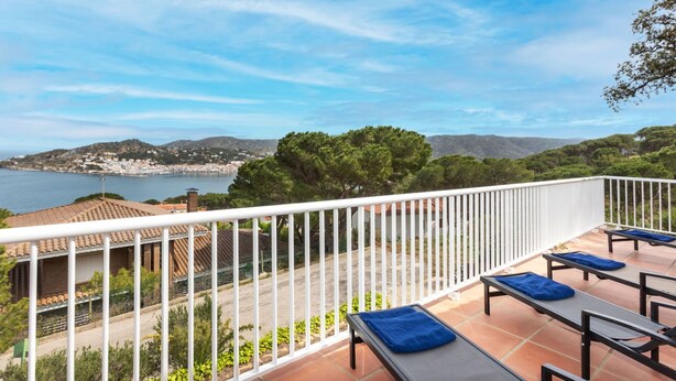 A terrace with outdoor seating and a scenic view of a coastal town and hills.