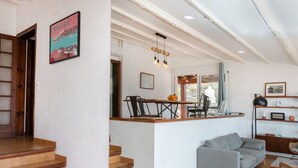 Amplia sala de estar con un sofá gris, una escalera de madera y una zona de cocina con mesa y sillas.