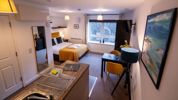 A hotel room with a bed, a desk, a chair, a window with a view of a stone building, and a painting on the wall.