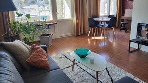 En moderne stue med sofa, lille bord og spiseområde med stole. Der er krukkeplanter og udsigt udefra gennem store vinduer.