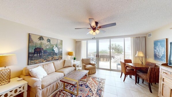 A spacious living area with a sofa, coffee table, and dining set. There is a ceiling fan, a large painting, and a view through the glass doors.