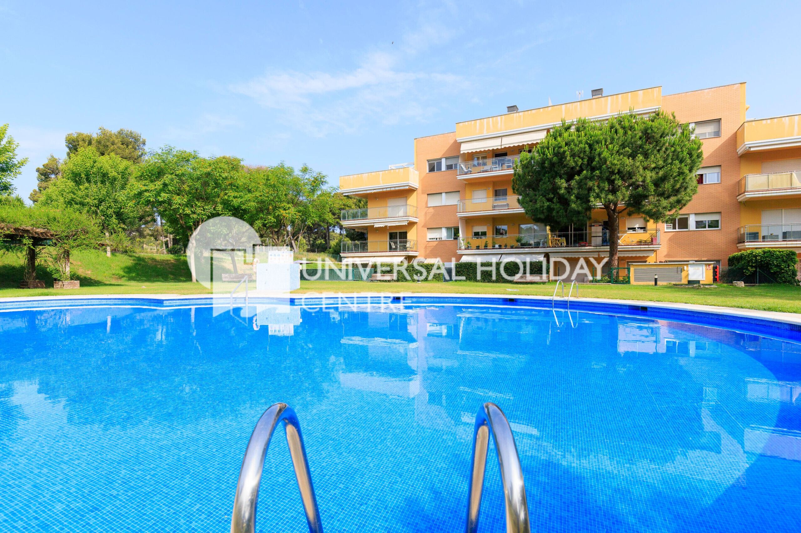 A swimming pool with a clear blue water surface, surrounded by a well-maintained lawn and trees. In the background, there is a multi-story building with balconies and a sign that reads "UNIVERSAL HOLIDAY".
