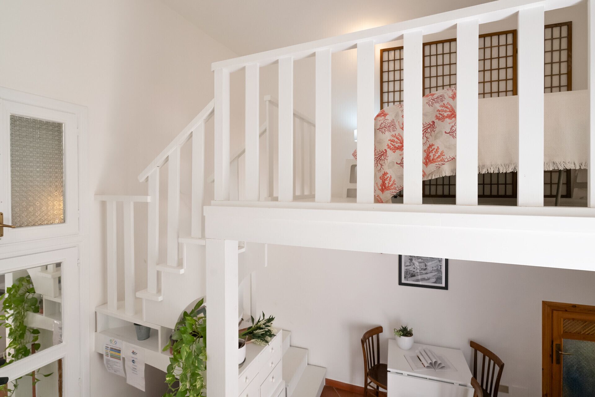 Uma escadaria branca com corrimão, uma área de refeições com mesa e cadeiras, e uma janela com um painel decorativo.