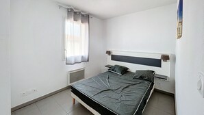 Una habitación pequeña y limpia con una cama individual, una ventana con cortinas translúcidas, un estante en la pared y una lámpara en el techo.