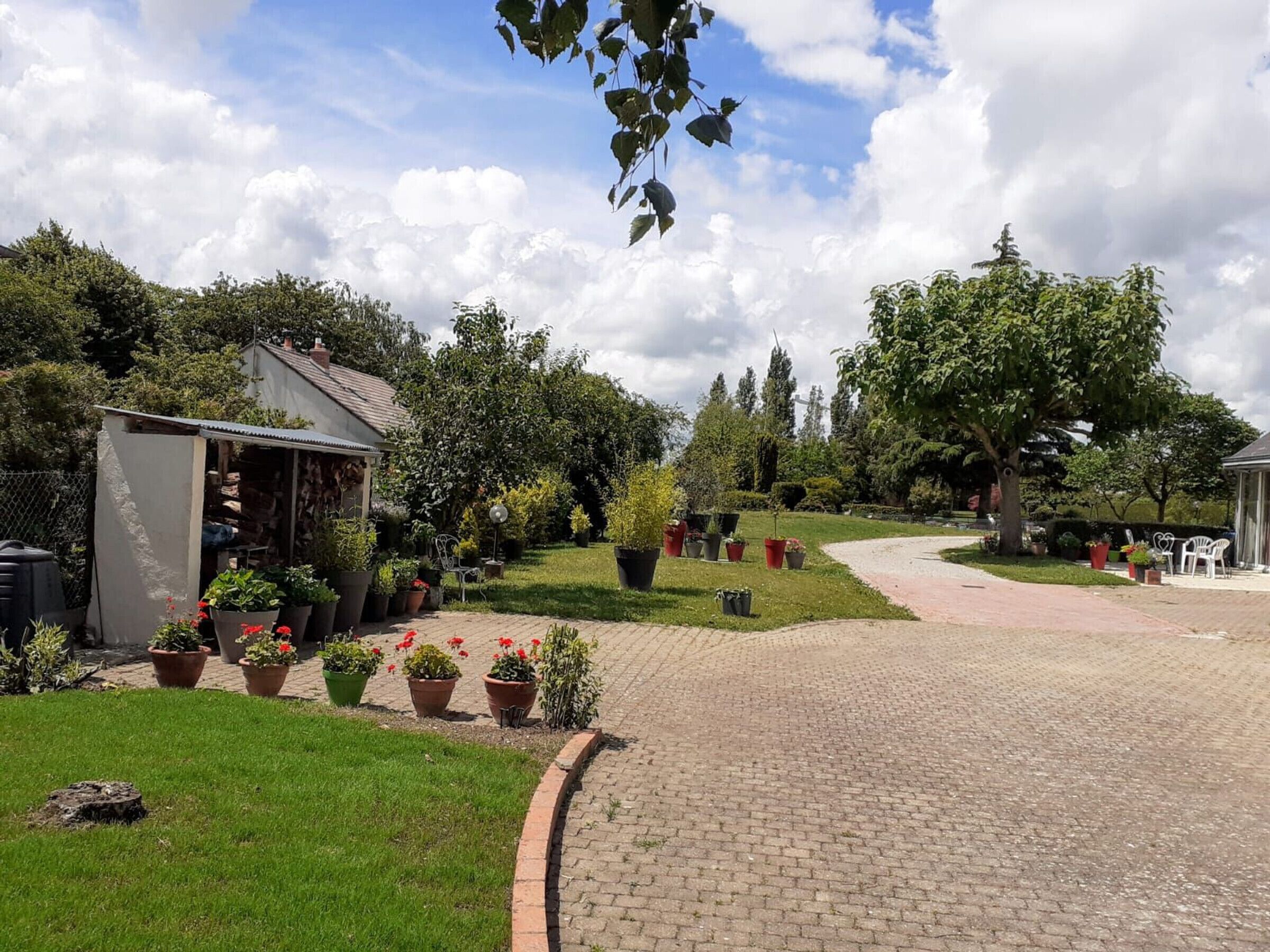 A paved pathway leading through a garden with potted plants and a small structure on the left.