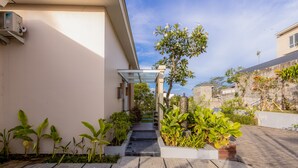 Un patio moderno al aire libre con cubierta de madera, plantas en macetas y un pasillo cubierto de vidrio que conduce a la entrada de un edificio.