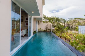 Un área moderna junto a la piscina con barandilla de vidrio, un pequeño rincón de descanso con una silla y una mesa, y vistas a la vegetación y edificios circundantes.