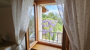 Una habitación con piso de madera, una ventana con cortinas blancas y vistas a flores moradas al exterior.