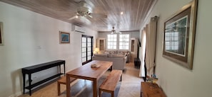 A living room with a sofa, a wooden table, and a bench. There are windows and a ceiling fan.
