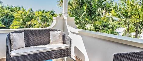 A balcony with wicker furniture, a glass table, and lush greenery in the background.