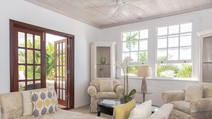Un amplio salón con sofá, sillones y una mesa de centro. Una puerta da a un área exterior con plantas.