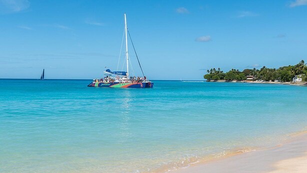 Una playa con agua turquesa cristalina, un velero y una costa lejana con edificios y árboles.