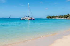 A beach with clear turquoise water, a sailboat, and a distant shoreline with buildings and trees.