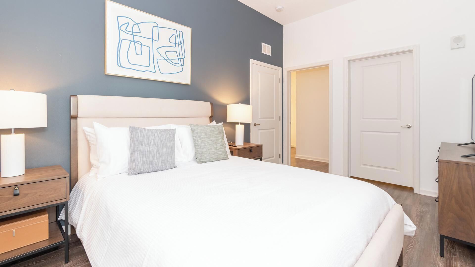 Un lit bien fait, avec du linge de lit blanc et deux oreillers, une table de chevet avec une lampe, et une œuvre d’art abstraite accrochée au mur.