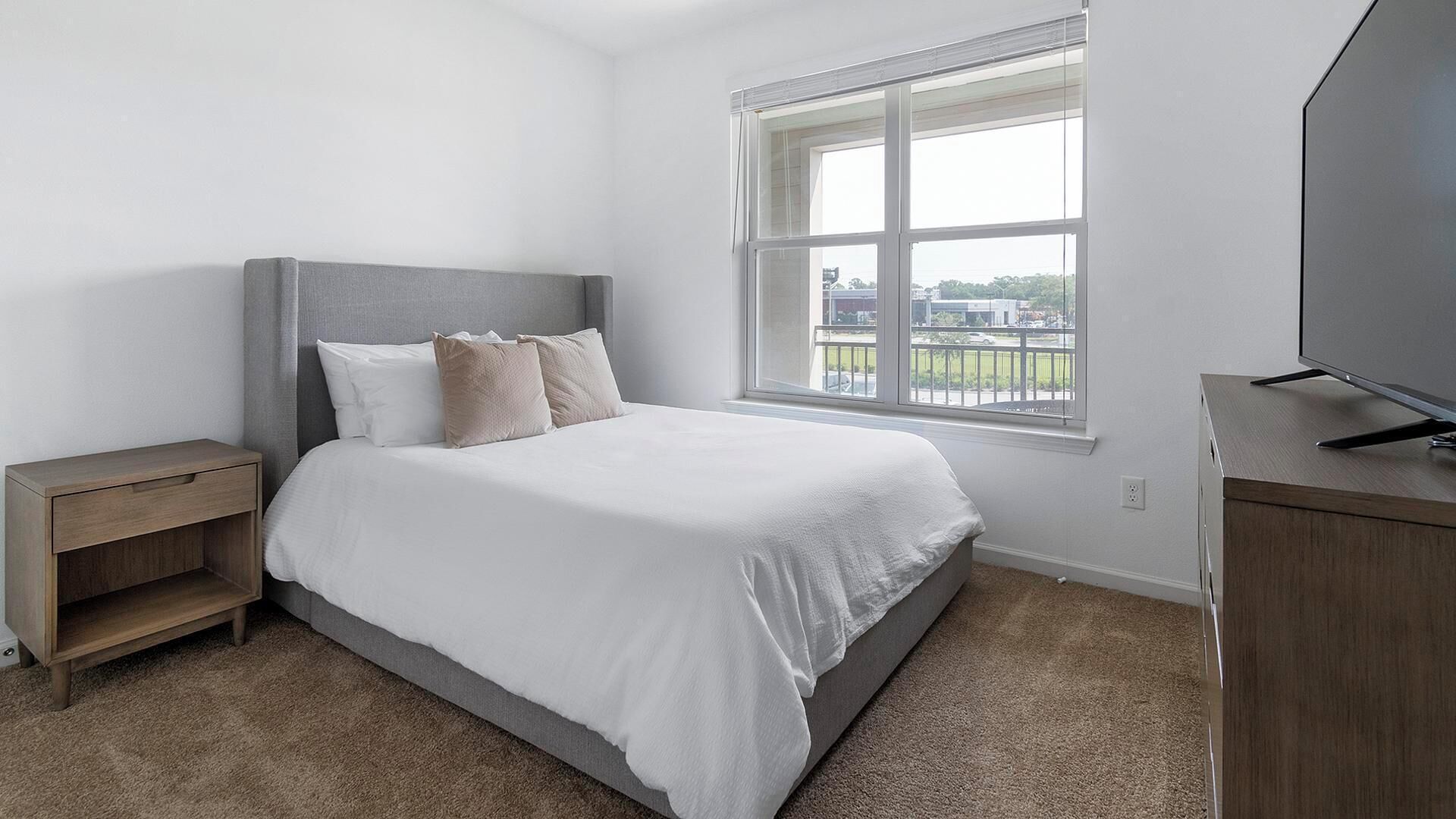 A bedroom with a bed, a nightstand, a TV, and a window with a view of buildings.