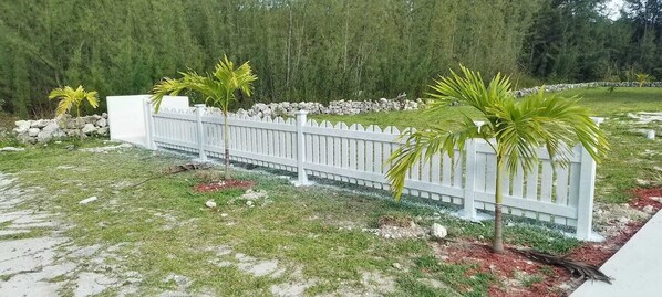 Una cerca blanca de piquetes con palmeras y un muro de piedra al fondo.