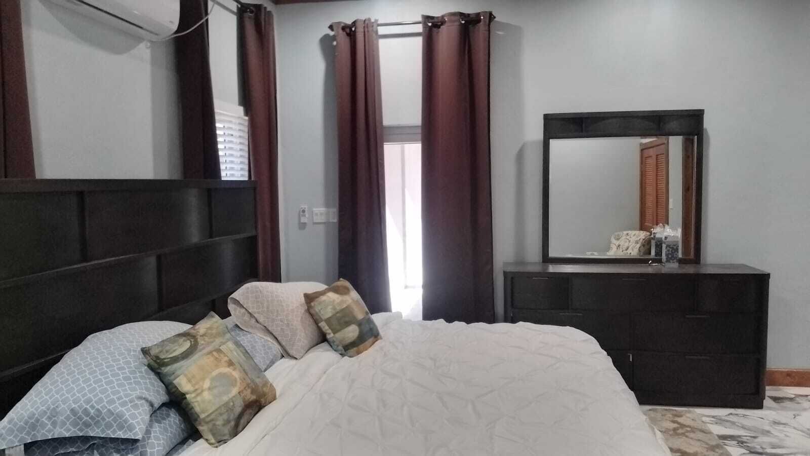 Een slaapkamer met een bed, een dressoir, een spiegel en een raam met gordijnen.