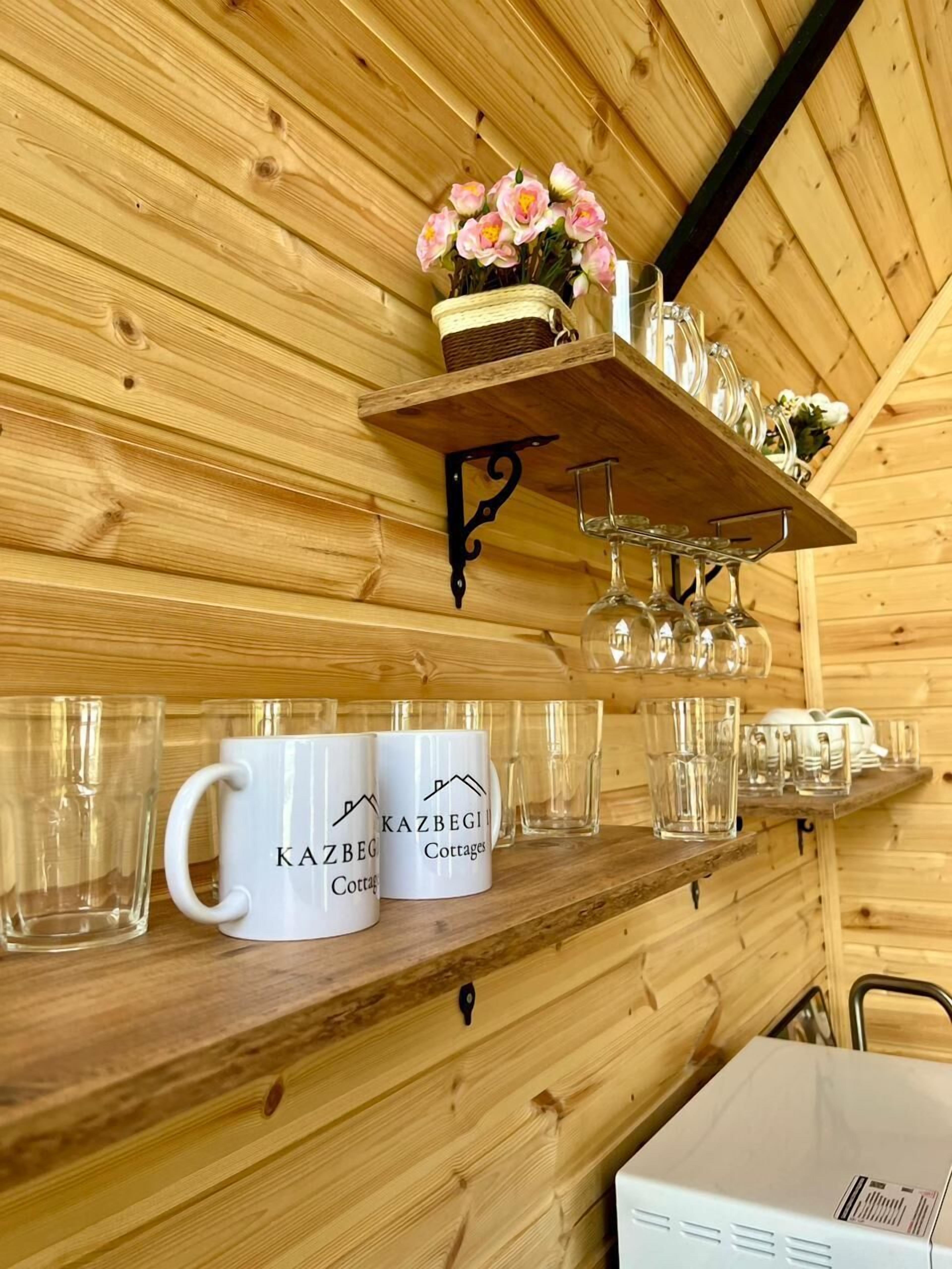 A wooden interior with a shelf holding mugs and glasses, a basket with flowers, and a sign reading "KAZBEGI Cottages".