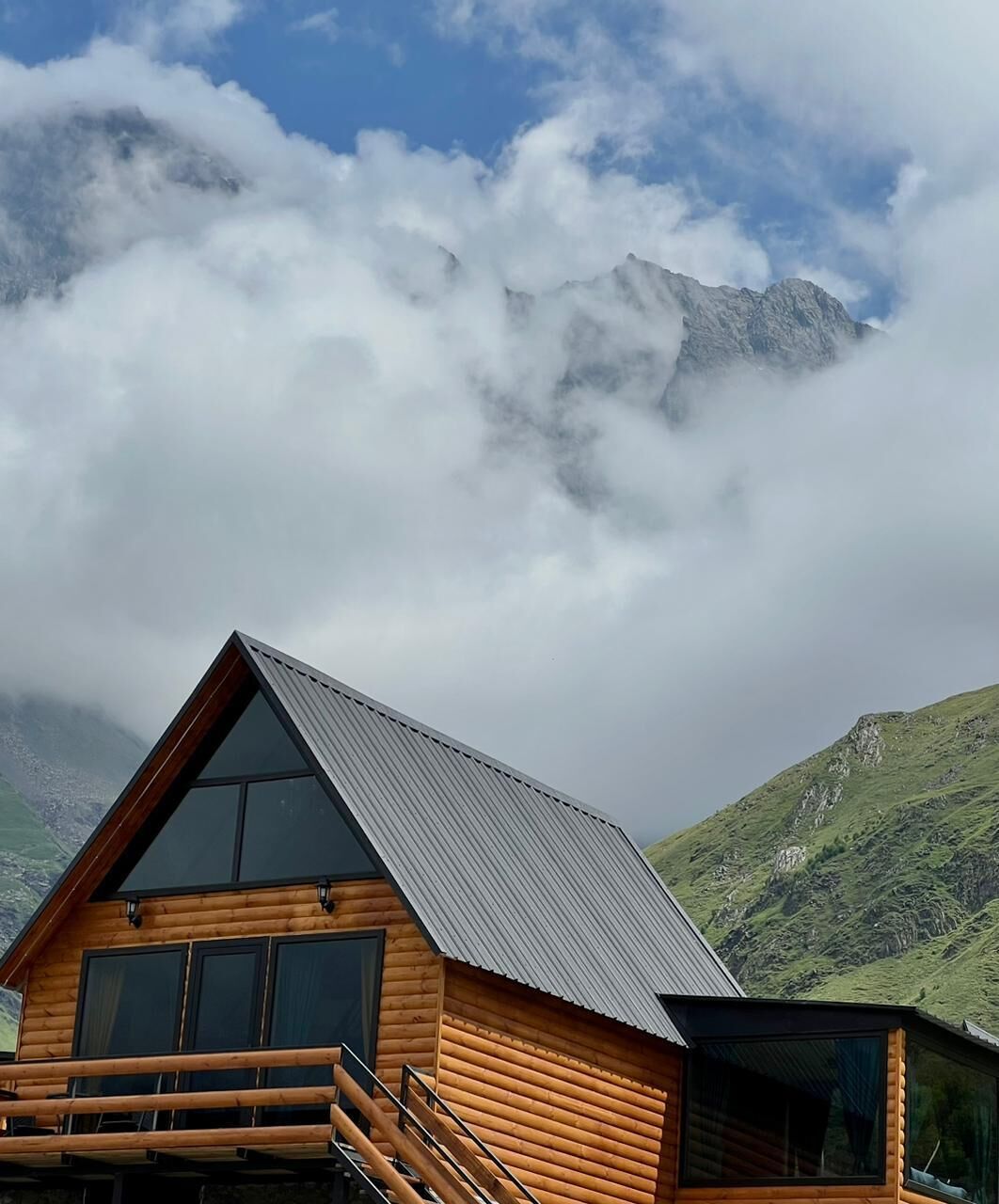 A wooden cabin with a sloped roof, large windows, and a balcony, set against a backdrop of mountains and clouds.